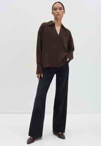 Femme debout avec une main dans la poche, portant un chemisier à manches longues marron foncé, un pantalon noir à jambes larges et des chaussures pointues marron.