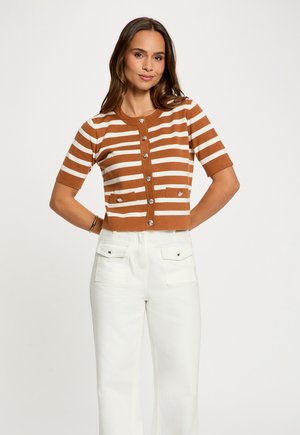 Kvinde iført en brun- og hvidstribet kortærmet cardigan med knapper og hvide bukser med høj talje, stående med hænderne bag ryggen.