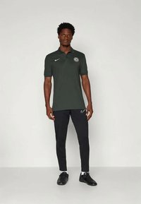 Mann steht in einem dunkelgrünen Nike-Polo mit Chelsea-Logo, schwarzen Nike-Hosen, weißen Socken und schwarzen Sneakers vor einem einfarbigen Hintergrund.