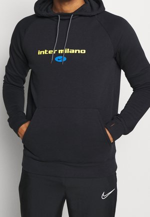 Mand iført sort hættetrøje med gul tekst "inter milano" og blåt Nike-logo, hænderne i frontlommen, kombineret med sorte Nike-bukser.