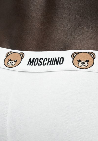 Ceinture de sous-vêtement blanche avec logo Moschino centré entre deux visages d'ours en peluche sur une peau foncée.