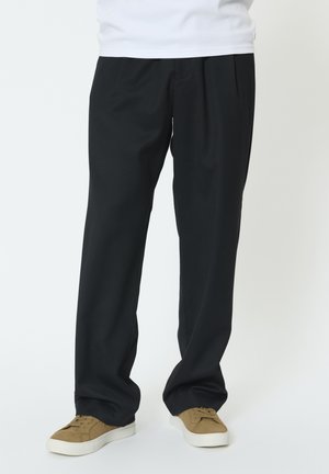 Osoba ubrana w luźne czarne spodnie i beżowe trampki z białymi podeszwami, stojąca na tle jednolitego jasnego tła.