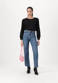 Svart langermet topp, blå denimjeans med perledekorasjoner i kanten, og svarte ankelstøvler; holder en liten rosa håndveske.