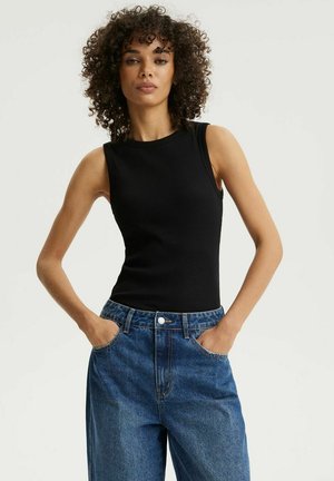 Kobieta o kręconych włosach, ubrana w czarny top bez rękawów i niebieskie, wysokie dżinsy, stojąca z rękami w kieszeniach na jednolitym tle.