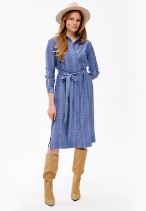 Kobieta nosząca niebieską sukienkę koszulową z wzorem i zawiązanym w talii paskiem, beżowe luźne kozaki do kolan oraz jasnobeżowy kapelusz typu fedora.