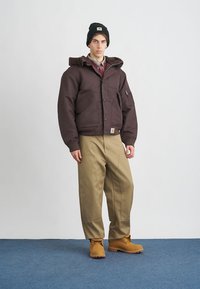 Braune isolierte Jacke mit Kapuze, khakifarbene Hose und braune Stiefel. Trägt dazu eine schwarze Mütze und ein mehrlagiges Oberteil. Eine einzelne Brusttasche ist sichtbar.