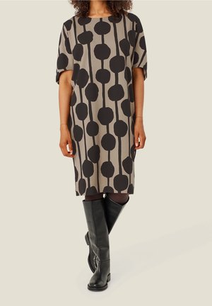 Femme portant une robe beige mi-longue avec des motifs verticaux de cercles noirs et des bottes en cuir noires montant jusqu'aux genoux.