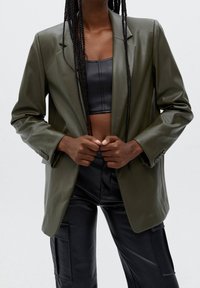 Blazer en cuir vert avec une silhouette ajustée, fermeture à un bouton et revers. Associé à un pantalon en cuir noir, mettant en valeur une texture lisse.