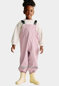 Un jeune enfant portant une salopette imperméable rose avec des boucles noires, un pull crème et des bottes de pluie jaunes, se tenant devant un fond uni.
