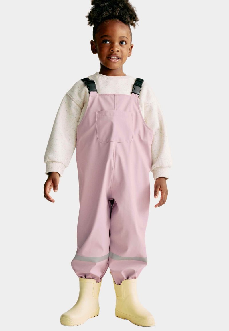 Un jeune enfant portant une salopette imperméable rose avec des boucles noires, un pull crème et des bottes de pluie jaunes, se tenant devant un fond uni.