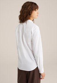 Camicia bianca a maniche lunghe con colletto classico, tessuto liscio e orlo curvo, abbinata a pantaloni marroni a righe.