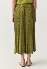 Grüne plissierte Midi-Rock mit einem strukturierten Zickzackdesign, elastischem Bund und Saum, der zu einer spitzen Form verjüngt, kombiniert mit Sandalen.