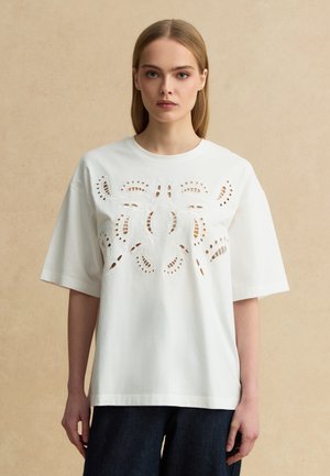 Junge Frau mit glatten blonden Haaren, die ein weißes übergroßes T-Shirt mit symmetrischem Blumen-Ausschnitt vorne trägt.