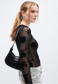 Μαύρη διάφανη μπλούζα με μακριά μανίκια που διαθέτει μοτίβα από βελούδο με λουλούδια, συνδυασμένη με μαύρη τσάντα ώμου με λεπτομέρεια πόρπης. Ορατό χρωματιστό παντελόνι ανοιχτού τόνου.