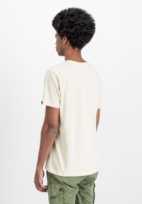 Kreem värvi lühikeste varrukatega T-särk, mis on valmistatud pehmest puuvillast. Ümmargune kaelus, tavaline lõike ja sildiga detail vasakul varrukal. Ükski muster ei ole nähtav.