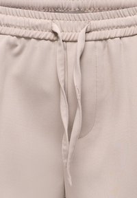Pantalon beige clair avec ceinture élastique, cordon en tissu noué à l'avant et détails de coutures visibles.