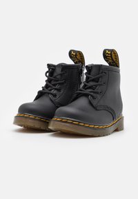 Bottes à chevilles en cuir noir avec coutures jaunes, devant à lacets et anses de tirage. Semelles en caoutchouc pour l'adhérence et la durabilité. Design compact et structuré.