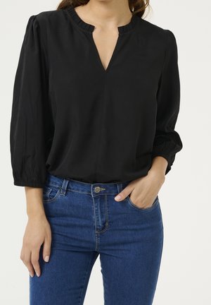 Kobieta w czarnej bluzce z długim rękawem i falbankowym kołnierzykiem oraz niebieskich dżinsach, z jedną ręką w kieszeni, na jednolitym jasnym tle.