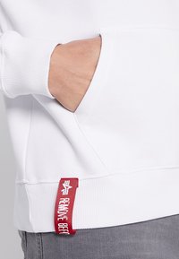 Hvit hettegenser med ribbestrikkede mansjetter, kengurulomme og en fremtredende rød etikett på siden med tekst og logo detaljer.
