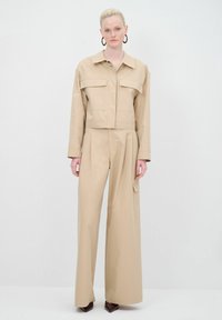 Giacca beige cropped con due tasche frontali, abbinata a pantaloni a gamba larga con una tasca laterale. Tessuto liscio con un design strutturato.