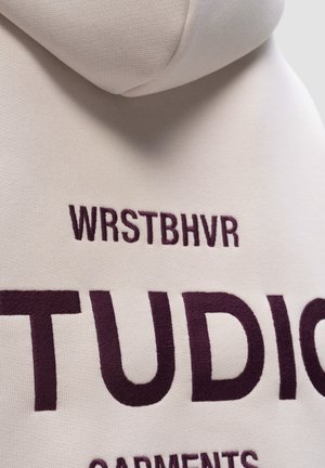 Beige hoodie met een gestructureerde afwerking. Heeft bordeauxrood geborduurde tekst: "WRSTBHVR" boven "STUDIO" en "GARMENTS" daaronder.