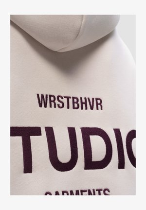 Beige hoodie met een gestructureerde afwerking. Heeft bordeauxrood geborduurde tekst: "WRSTBHVR" boven "STUDIO" en "GARMENTS" daaronder.