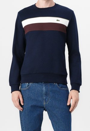 Homme portant un sweat-shirt bleu marine avec des rayures horizontales blanches et bordeaux sur la poitrine et un jean bleu, debout, les mains détendues.