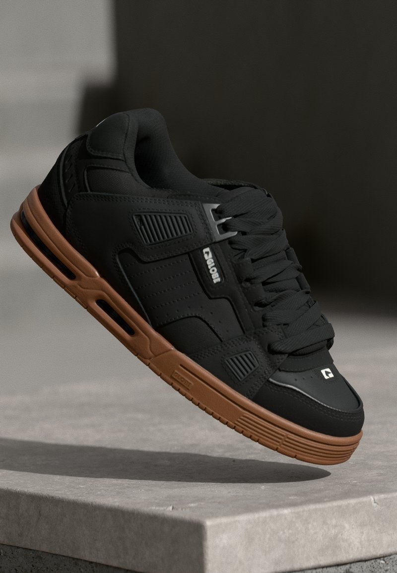 Sort sneaker med en glat læderoverdel, perforerede detaljer og brune gummisåler. Har rund tå, flade snørebånd og logodetalje på siden.