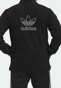 Svart fleecejacka med en vit Adidas-logotyp på ryggen, med dragkedja i kragen och långa ärmar. Kombinerad med svarta byxor och ränder.