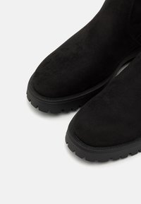 Botas de ante negras con punta redondeada y suela texturizada, que presentan un borde liso y un patrón de relieve prominente.