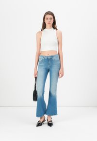 Weißes geripptes Crop-Top, hellblaue Schlagjeans mit ausgewaschenem Effekt und schwarze spitz zulaufende Schuhe. Schwarze Schultertasche. Sanfte Texturen.