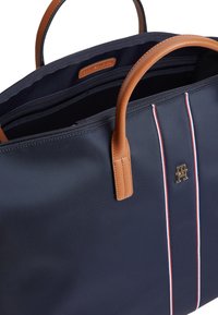 Τσάντα ώμου από νάιλον σε ναυτικό μπλε χρώμα με καφέ δερμάτινες λαβές, που διαθέτει δύο ρίγες σε κόκκινο και λευκό και χρυσό λογότυπο στο μπροστινό μέρος. Κλείσιμο με φερμουάρ στην κορυφή.