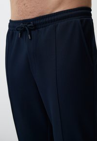Gros plan sur une personne portant un pantalon en cordon de serrage bleu marine foncé avec une taille élastique et des poches latérales, montrant le torse et le haut des jambes.