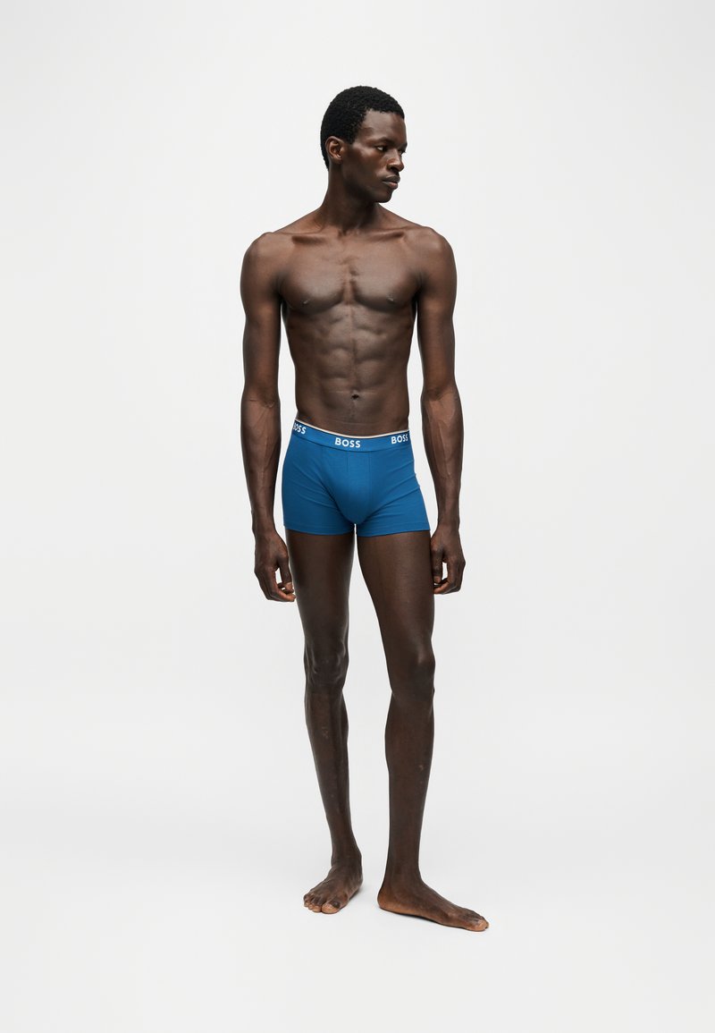 Lange, slanke zwarte man staat op blote voeten en draagt blauwe BOSS boxershorts, kijkend naar links tegen een effen witte achtergrond.