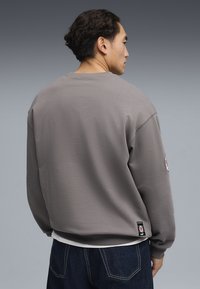 Sudadera gris de corte holgado, con cuello redondo y puños acanalados. Presenta un parche con el logo en la manga y detalles de costuras en contraste.