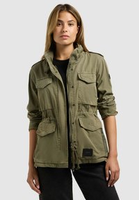 Mujer de cabello largo que lleva una chaqueta de estilo militar verde oliva con múltiples bolsillos y mangas remangadas, de pie contra un fondo liso.