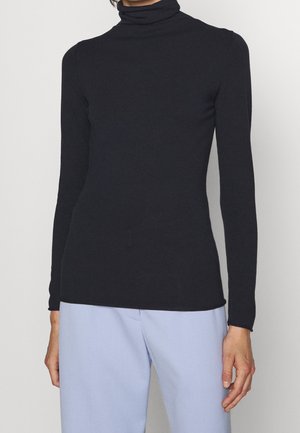 Personne portant un pull noir moulant à col roulé à manches longues associé à un pantalon taille haute bleu clair, sur un fond uni.