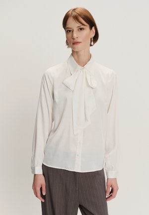 Weiße Bluse mit einer großen Schleife am Hals, langen Ärmeln, Knopfverschluss und glatter Textur, kombiniert mit grauen Nadelstreifenhosen.