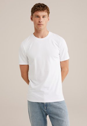 Junger Mann mit hellbraunem Haar, der ein schlichtes weißes T-Shirt mit kurzen Ärmeln und hellblaue Jeans trägt, vor einem beigen Hintergrund.