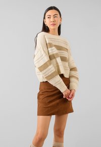 Maglione corto a righe crema e marrone con una vestibilità morbida, abbinato a una mini gonna in suede marrone. Texture lavorata a maglia e polsini a costine visibili.