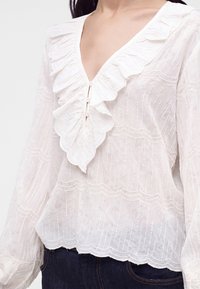 Blouse blanche transparente avec col en V à volants, détails de boutons, ourlet festonné et manches longues portée avec un jean foncé.