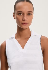 Bela brezrokavna polo majica z V-izrezom, strukturiranimi črtami in kontrastnim obrobjem ob izrezih rokavov. Gladek zaključek tkanine, minimalen dizajn.