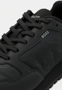 Zapatilla deportiva negra con una parte superior texturizada, acentos lisos, cordones planos y un logotipo prominente. Suela con dibujo para tracción.
