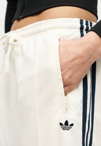 Κοντινό πλάνο ενός ατόμου που φοράει λευκό παντελόνι φόρμας Adidas με μπλε ρίγες, ελαστική μέση και το χέρι του τσουγκρισμένο στην πλαϊνή τσέπη.
