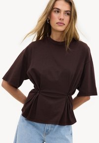 Jeune femme portant un haut marron foncé à manches courtes avec des détails froncés à la taille et un jean bleu clair, posant devant un fond blanc.