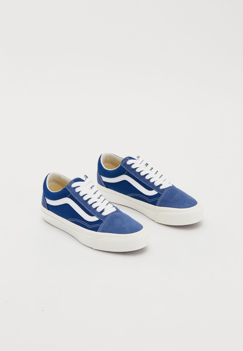 Paire de baskets basses bleues et blanches avec des lacets blancs et des semelles en caoutchouc sur un fond blanc uni.