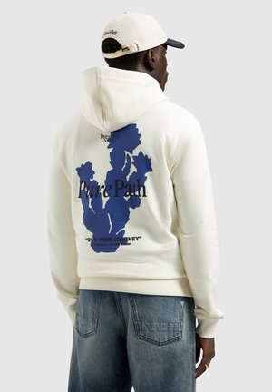 Man draagt een crème-kleurige hoodie met een blauwe abstracte cactus en de tekst "Pure Path" en "Own Your Journey", gecombineerd met lichtblauwe jeans en een witte pet.