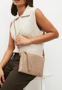 Trblietavý béžový crossbody vak so zipsovým uzáverom. Vyrobený z textúrovaného materiálu, obsahuje odnímateľný popruh a bočný detail s strapcom.