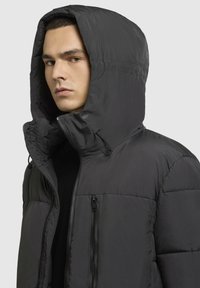 Mand iført en sort polstret hættejakke med lynlås foran og brystlomme, som vender let til venstre på en lys ensfarvet baggrund.