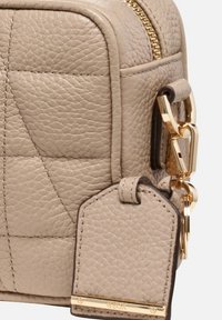Borsa a mano in pelle beige con superficie testurizzata, design trapuntato, hardware dorato e un'etichetta staccabile con logo e accento dorato.